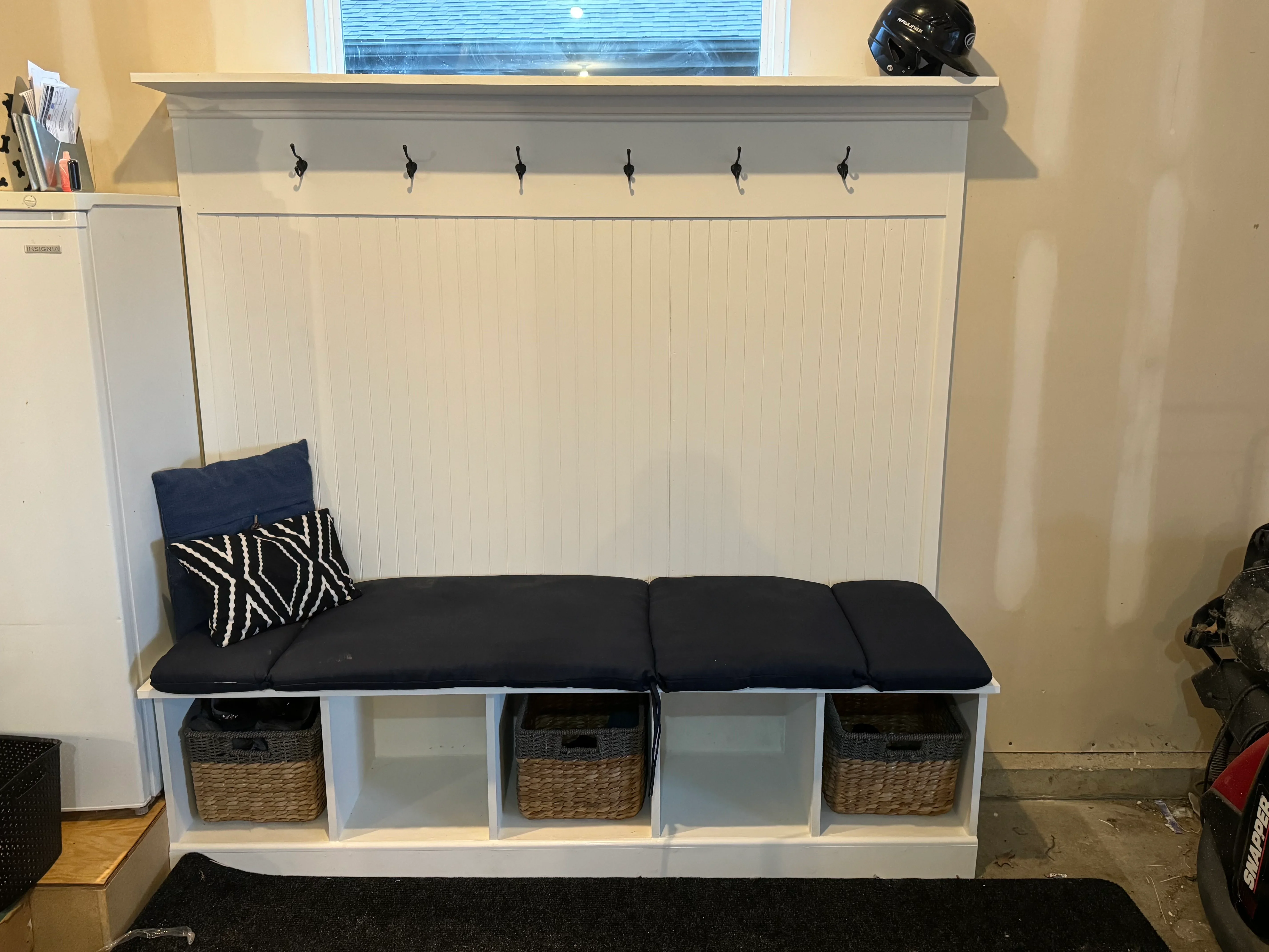 White built-in mudroom bench with black cushion, storage cubbies, woven baskets, and six wall hooks