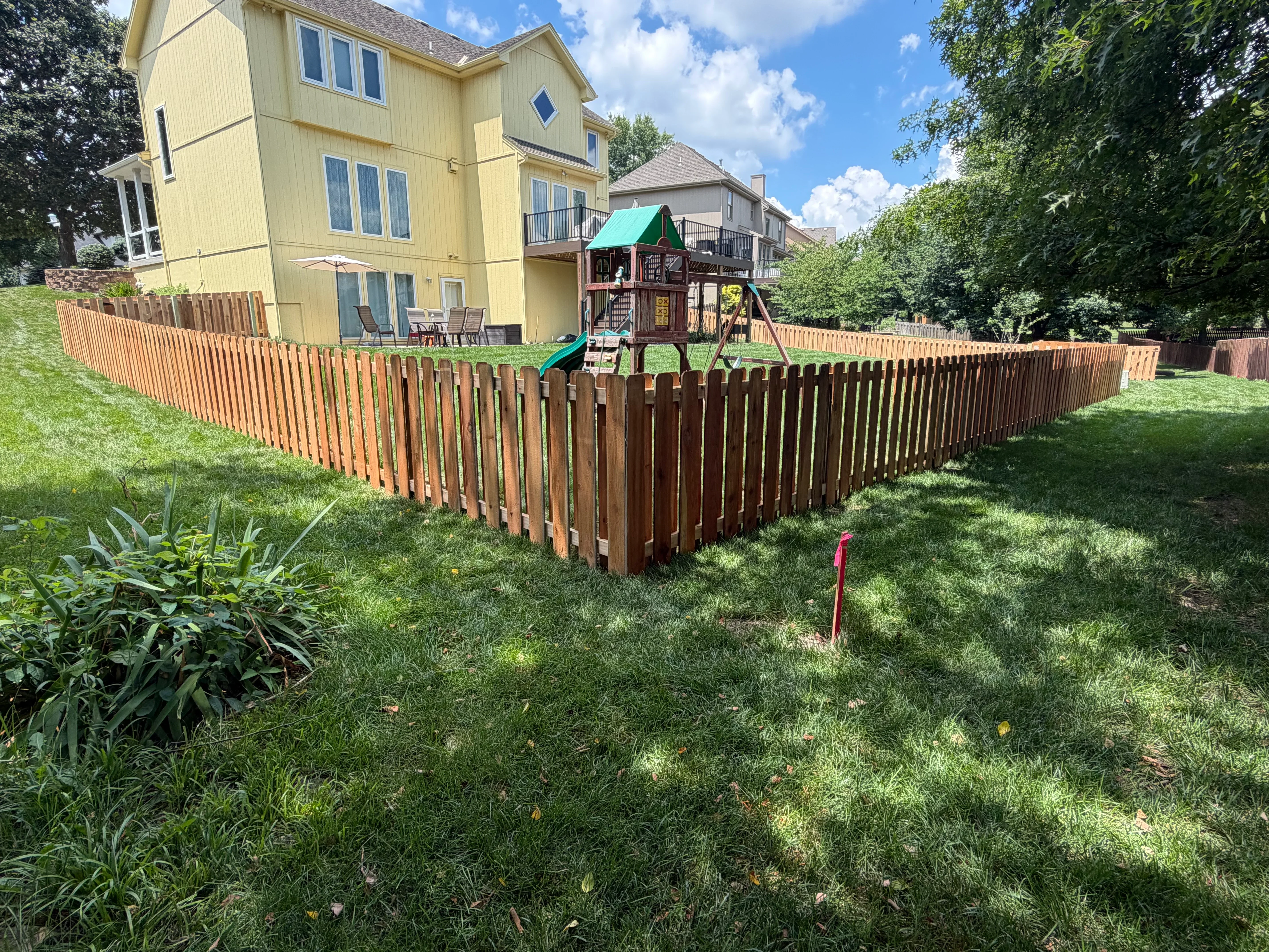 Stained wood picket fence surrounding backyard with play structure and mature landscaping